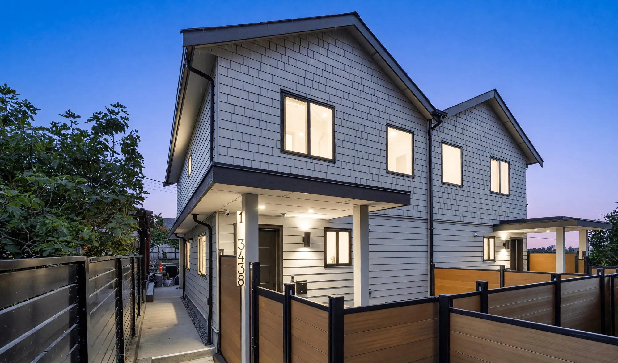 1-3438 Triumph Street at dusk: a new two-storey house with lit windows behind a timber and steel fence, reached along a private walk